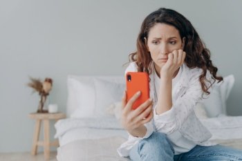 Stressed caucasian girl is reading morning news looking at smartphone display. Young woman is chatting on phone or browsing internet at home in her bedroom. Lady is doom scrolling.. Stressed caucasian woman is doom scrolling. Girl reading morning news on smartphone in her bedroom.