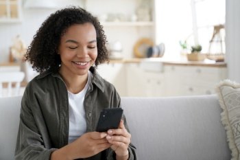 Happy african american teen girl share good news on social network via mobile phone sitting on sofa at home. Smiling mixed race young female enjoying shopping online using smartphone apps.. Happy african american teen girl holds smartphone using social network apps, shopping online at home