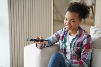 Happy african american young girl holding using tv controller, sitting on couch in living room, watching movie or series at home. Smiling teen lady relax at home spending leisure time with television.. African american young girl watching movie or tv series, enjoying television program at home