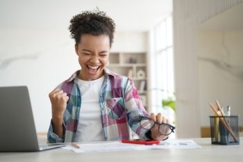 Excited happy african american high school girl, celebrating victory, got email with good exam test scores. Overjoyed biracial teen student got good news online sitting at laptop. Distance education. Excited biracial girl student celebrates victory, got email with good scores. Personal achievement