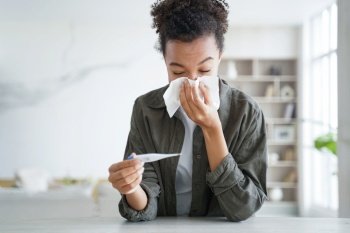 Teenage afro girl caught cold. Young hispanic woman is measuring temperature with thermometer, sneezing and blowing running nose. Flu or coronavirus symptoms. Girl is staying home while sick.. Young hispanic woman is measuring temperature with thermometer, sneezing and blowing running nose.