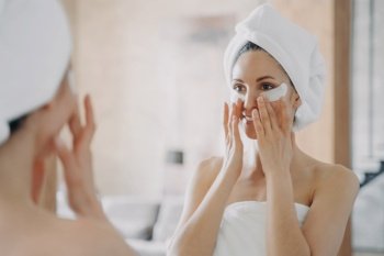 Girl applies eye patches and looking into mirror. Attractive european woman wrapped in towel after bathing. Young hispanic lady takes shower at home. Relaxation at spa resort. Beauty routine at home.. Girl applies eye patches and looking into mirror. Attractive woman wrapped in towel after bathing.