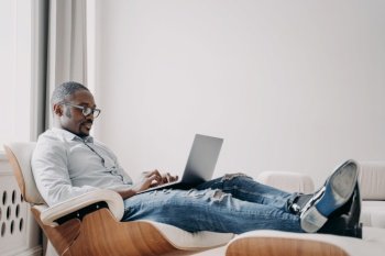 Focused african american man freelancer working online at laptop, typing email or message, sitting in modern comfortable armchair. Black male businessperson developing business project. Remote work. African american man working online on business project at laptop, sitting in armchair. Remote job