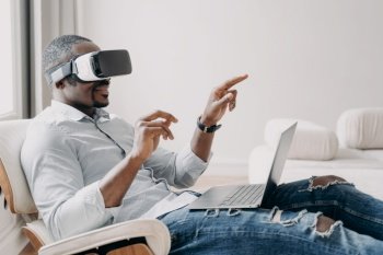 Relaxed businessman clicks virtual buttons. Gestures of african american man in vr glasses. Guy is working at home. Distant work and futuristic gaming. Modern digital technology for business.. Relaxed businessman clicks virtual buttons. Gestures of african american man in vr glasses.