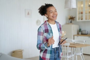 Excited happy girl in airpods is holding mobile phone. Teenage african american girl is singing song which is listening in wireless earphones. Weekend morning at home. Music service website concept.. Excited happy girl in airpods is holding mobile phone. Teenage girl is singing in weekend morning.