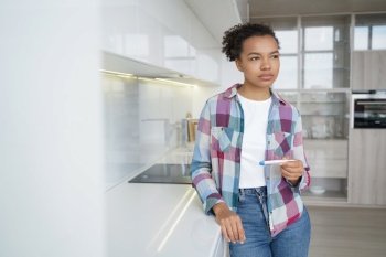 Pensive teenage girl gets positive pregnancy test result. Sad african american young woman is holding stripe test. Teenage lifestyle, unwanted pregnancy and motherhood. Contraception concept.. Pensive teenage girl gets positive pregnancy test result. Unwanted pregnancy and motherhood.