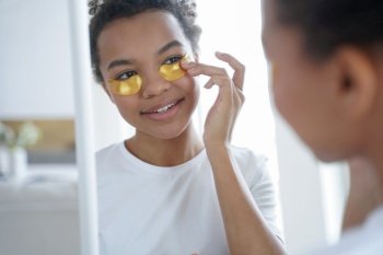 Young teen biracial girl applying moisturizing under eye patches look at mirror. Happy african american woman using gold hydrogel pads reducing facial puffiness, enjoy daily skincare routine.. Young teen african american girl applying moisturizing under eye patches, looks at mirror. Skincare