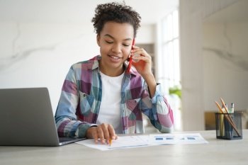 Young mixed race teen girl discussing homework with classmate by phone sitting at desk with laptop. Friendly schoolgirl student chatting with friend during exam preparation at home.. Biracial teen girl call by phone, doing homework, sitting at desk with laptop. Education and tech