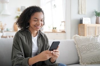 Curly african american girl has fun with phone at home. Happy hispanic woman is browsing social media and smiling. Chatting with friends remotely. Messaging with classmates. Communication concept.. Curly african american girl has fun with phone at home and smiling. Communication concept.