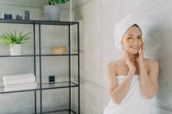 Relaxation at spa resort. Happy girl wrapped with her hair and body in towels applies eye patches. Young hispanic lady takes shower at home. Face skin rejuvenation and collagen nourishing.. Relaxation at spa resort. Happy girl wrapped with her hair and body in towels applies eye patches.