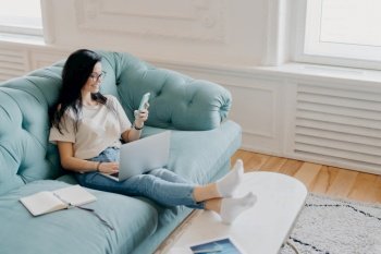 Horizontal shot of woman freelancer earns money online, checks email on smartphone, surrounded by modern technologies, organizer for making notes, uses high speed internet, sits on comfortable couch
