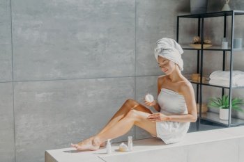 Female beauty and skin care concept. Young pretty woman applies cream, has slender legs, makes cosmetological procedure, wrapped in towel, feels perfect, takes care of her body and appearance