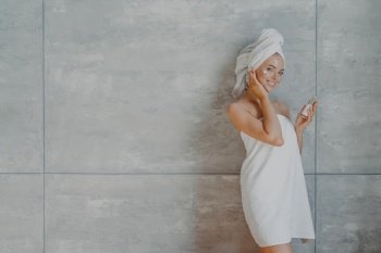 Happy healthy young woman with tender facial skin, applies beauty cream and undergoes beauty treatments, wrapped in bath towel, poses against grey background with copy space for your promotion