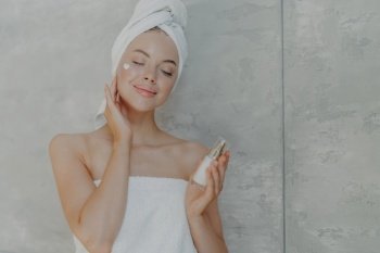 Pretty young woman holds bottle of body lotion, applies face cream on complexion, stands with closed eyes, wears bath towel on head, poses against grey wall. Getting pleasure from skin pampering