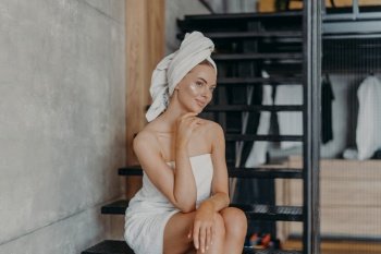 Thoughtful young European woman keeps hand under chin, sits on stairs and has thoughtful expression, has healthy skin, puts face cream, wrapped in bath towel, undergoes beauty treatments at home