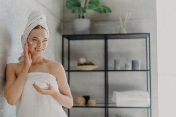 Lovely smiling Caucasian woman applies beauty cream on cheek, enjoys morning domestic skin care routine, wrapped in bath towel, grooming herself after showering poses in bathroom. Hygiene concept