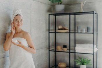 Photo of relaxed young woman applies face cream, has satisfied expression, wrapped in bath towel, poses near grey wall in bathroom, uses cosmetic product for rejuvenation. Beauty and health care