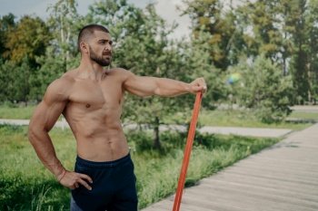 Muscular athletic man has workout with elastic resistance band poses with shirtless muscular body, stands outdoor, concentrated into distance. Athlete sportsman has workout in park. Healthy lifestyle