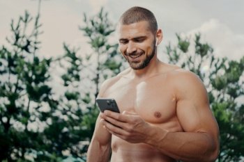Cheerful motivated shirtless sportsman concentrated in smartphone, sends text messages, uses wireless earphones for listening music, has strong muscles, stands outdoor. Sporty lifestyle concept