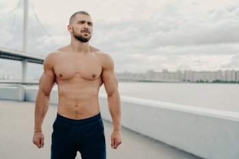 Young bearded athletic man with naked torso muscular body looks somewhere into distance, dressed in sport shorts, rests during workout, poses outdoor near river bridge. Fitness workout concept
