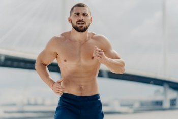 Half length shot of serious male jogger runs actively outdoor concentrated into distance poses with naked torso poses against blurred background with bridge. Fit muscular man runner trains actively