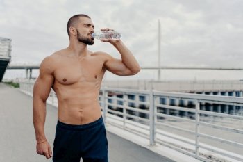 Active sportsman drinks bottle of water poses with naked torso concentrated into distance feels thirsty after running outdoors being full of energy, leads active lifestyle. Sport for better life