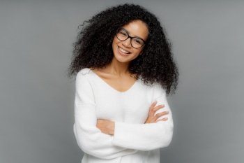 Half length shot of friendly looking African American woman tilts head, has dark crisp bushy hair, keeps arms folded over chest, wears casual white sweater, enjoys photoshoot in studio. Emotions