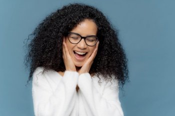 Overemotive female touches cheeks laughs from joy, keeps eyes closed, cannot stop giggling, has Afro hairstyle, dressed in white jumper, models against blue background. Ethnicity and emotions concept