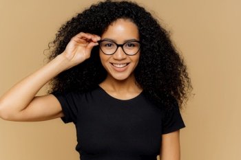 Lovely young African American woman grins at camera, keeps hand on rim of spectacles, glad to hear good news, dressed in casual black t shirt, isolated over brown background. Happiness concept