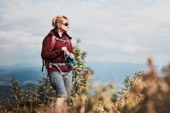 Woman with backpack hiking in mountains, spending summer vacation close to nature. Woman walking on top of hill admiring mountain landscape panorama holding bottle with water