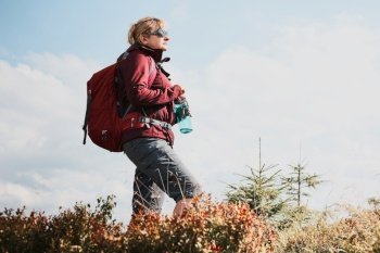 Woman with backpack hiking in mountains, spending summer vacation close to nature. Woman standing on top of hill admiring mountain landscape panorama holding bottle with water