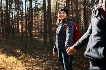 Couple holding hands enjoying trip while vacation day. Hikers with backpacks walking on forest path on sunny day. Active leisure time close to nature
