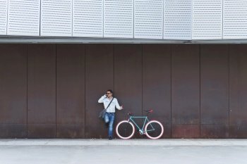 Young male with sunglasses and shoulder bag standing nest to a fixed gear bike while calling by phone