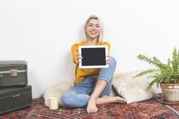 Portrait of a nice-looking attractive lovely charming cheerful cheery blonde haired girl holding in hands demonstrating new cool device while sitting on floor