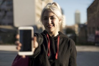 Girl standing against the city holding out a blank mobile phone towards the camera with focus to the person