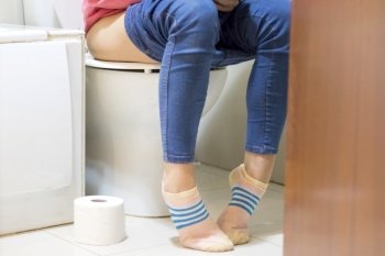 close up photo woman with constipation or diarrhea sitting on toilet with her pants down around her legs and a paper roll on the floor