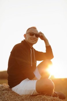 handsome young man in sunglasses, casual black sweatshirt and white short, in summer field outdoor, sitting on hay bale, haystack on sunset.. handsome young man in sunglasses, casual black sweatshirt and white short, in summer field outdoor, sitting on hay bale, haystack on sunset