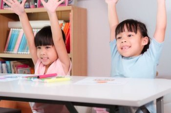 Happy kids with arms up sit in classroom. Cute girl taking lessons for home schooling Elementary school children enjoy learning together. Back to school concept.