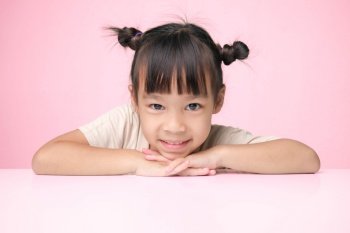 Cute dark hair little girl smiles happily and puts her hand under her chin while sitting at a table on a pink background and looking at the camera. Advertising childrens products