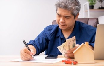 Unhappy Asian man holding a banknote and Making an account of income and expenses to pay the home loan and car loan, Concept loan payment and home insurance, economic problems, and inflation