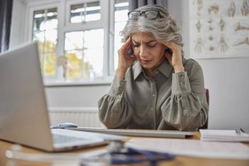 Stressed And Overworked Mature Female GP At Desk In Doctors Office
