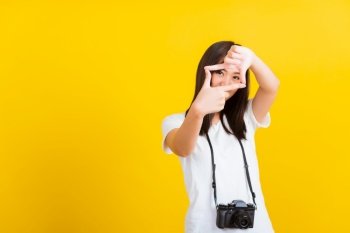 Portrait of happy Asian beautiful young woman photographer execute by hanging digital mirrorless photo camera on neck and show hand sign mark frame for photography, isolated on yellow background