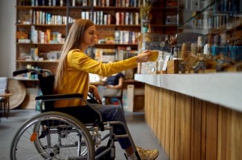 Disabled female student in wheelchair in cafe, disability, bookshelf and university library interior on background. Handicapped woman in college, paralyzed people get knowledge. Disabled female student in wheelchair in cafe