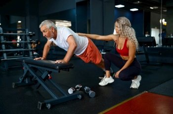 Old man doing push-up exercise on bench, female personal trainer, gym interior on background. Sportive grandpa with woman instructor in sport center. Old man, push-up exercise on bench, female trainer