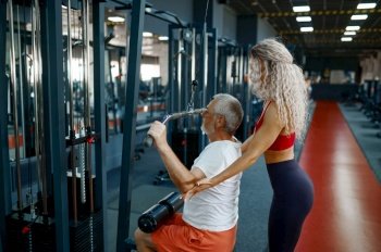 Old man on exercise machine, female personal trainer, gym interior on background. Sportive grandpa with woman instructor in sport center. Healty lifestyle, health care. Old man on exercise machine, female trainer, gym