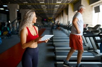 Old man on treadmill, female personal trainer with laptop, gym interior on background. Sportive grandpa with woman instructor, workout in sport center. Healty lifestyle, health care. Old man on treadmill, female trainer with laptop