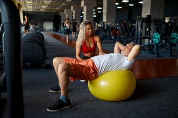 Old man doing exercise with ball, female personal trainer, gym interior on background. Sportive grandpa with woman instructor, fitness training in sport center. Healty lifestyle, health care. Old man, exercise with ball, female trainer, gym