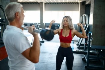 Old man doing exercise with bar, female personal trainer, gym interior on background. Sportive grandpa with woman instructor, workout in sport center. Old man doing exercise with bar, female trainer