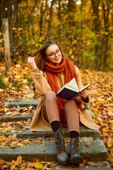 Closeup autumn portrait of young smart woman in eyeglasses and warm clothes reading book outdoors. Closeup of young woman reading book outdoors