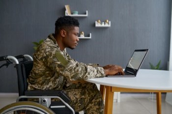 Military man with disability sitting in wheelchair working online using laptop computer. Military man with disability working on laptop